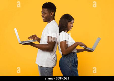 Couple Works Together on Laptops While Standing Back to Back Against a Bright Yellow Background Stock Photo