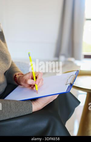 Close up of a woman writing on a clipboard with a pencil in a bright interior. Concept of planning, coaching, therapy session, notes and focused work. Stock Photo