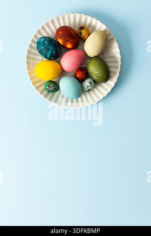 A variety of decorated Easter eggs in vibrant colors are creatively arranged on a white plate set against a soft blue background, symbolizing spring f Stock Photo
