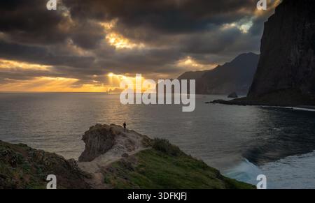 Golden sunbeams break through heavy clouds over the Atlantic Ocean near Miradouro do Guindaste while a solitary runner moves along a cliff edge Stock Photo