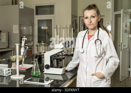 Young, Caucasian white, female laboratory technician in white robe with stethoscope standing next to compound microscope, laboratory, equipment and gl Stock Photo