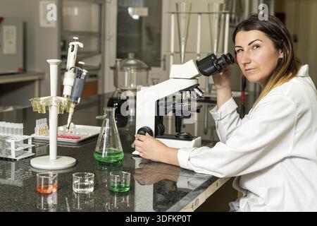 Middle age, Caucasian white, female laboratory technician in white robe with stethoscope sitting next to compound microscope, laboratory, equipment an Stock Photo