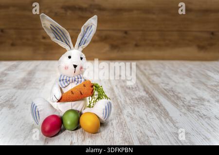 Close-up of three colorful Easter eggs in front of Easter bunny holding carrot on white painted and vintage oak background, front view Stock Photo