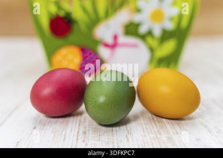 Close-up of three colorful Easter eggs in front of blurred felt Easter basket on white painted and vintage oak background, front view Stock Photo