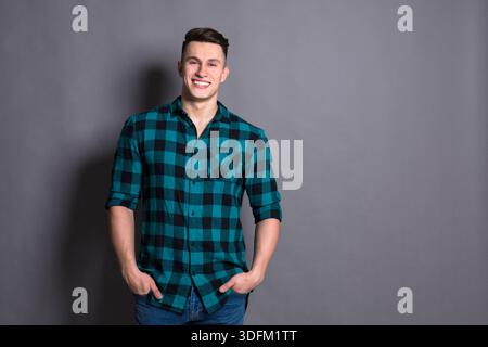 Young Man Smiling While Standing Against a Gray Backdrop With Hands in Pockets Stock Photo