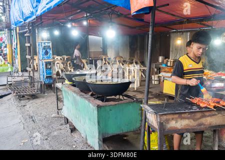 Young Man vendor is grilling food skewers and preparing other dishes in large woks, busy, open-air, street food stall, Cebu city, Philippines Stock Photo