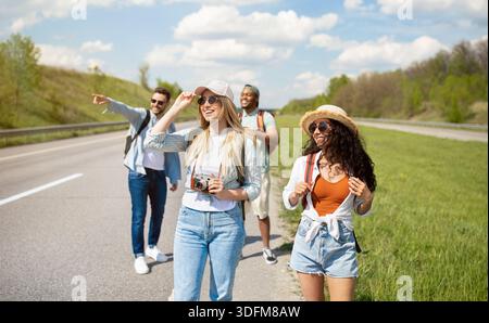Group of millennial multiracial friends with backpacks walking along road, traveling together outdoors Stock Photo