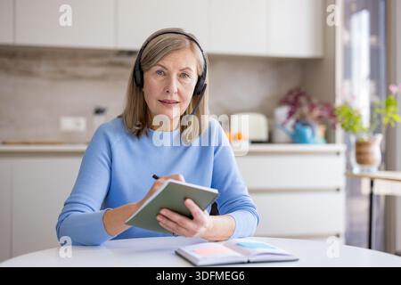 Senior woman wearing a headset and holding a notebook with a pen, actively participating in a virtual conference or remote work session from her home office setup in the kitchen Stock Photo