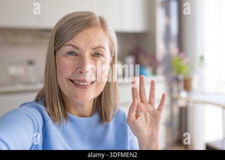 Senior woman looking at camera and waving hand, making a video call or taking a selfie in her home kitchen, connecting with family and friends through modern technology Stock Photo