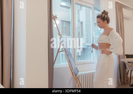 Woman stands before canvas near window, white outfit, brush raised, cool outdoor backdrop visible through glass, focused creative stance. Stock Photo