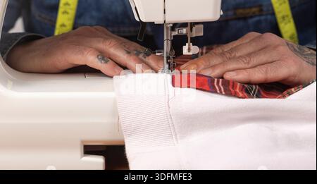 Person's tattooed hands guiding fabric under a sewing machine's needle, creating clothing Stock Photo