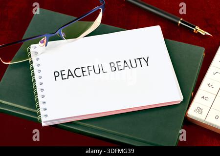 A spiral notebook with the words Peaceful Beauty is on a closed, green book. Eyeglasses, a pen, and a calculator are nearby on a dark red surface. Stock Photo