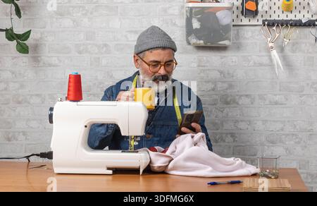 Man using mobile phone and holding a mug, sitting at sewing machine in a workshop Stock Photo