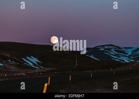 View of a mesmerizing full moon hangs in the dusk sky over dark, rolling hills touched with snow, above a stark road lined with yellow markers, Saudar Stock Photo