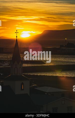 View of the sun setting behind a church steeple and distant mountains, casting warm golden hues across the tranquil waters, Saudarkrokur, Skagafjordur Stock Photo