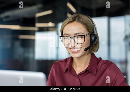 Smiling woman wearing a headset and eyeglasses providing professional customer support service, having a friendly conversation during a video call in a modern office Stock Photo