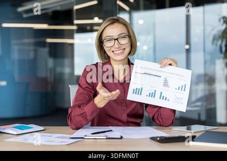Professional young businesswoman showing a financial data report with bar charts and graphs, discussing business analytics during a presentation in a modern office meeting Stock Photo