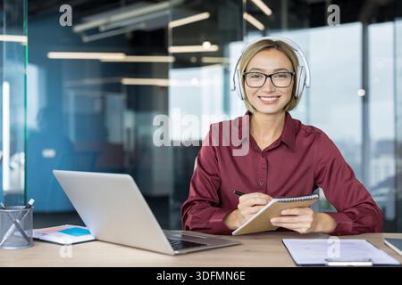 Young professional businesswoman in glasses and headphones smiles at camera while taking notes at a laptop on her office desk, confident and engaged in remote work or meeting Stock Photo