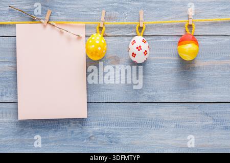 Colorful Decorative Eggs Hanging on Yellow String Above Blank Note on Wooden Background Stock Photo