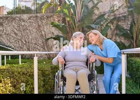 A senior woman in a wheelchair is assisted by a physiotherapist outdoors in a rehab center. This image represents recovery and rehabilitation after in Stock Photo