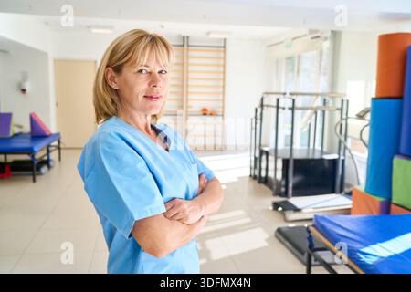 A confident physiotherapist stands with arms crossed in a rehabilitation center The medical professional wearing a blue uniform offers healthcare and Stock Photo