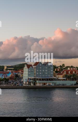 View of colorful buildings lining the waterfront, illuminated by the soft glow of the setting sun contrasting with the dark water, Willemstad, Curaçao Stock Photo