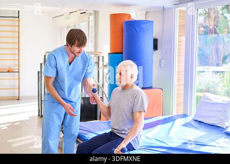 A senior man is doing arm exercises with dumbbells assisted by a physiotherapist in a rehab center. This image can be used for commercial purposes rel Stock Photo
