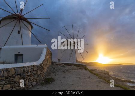 View of whitewashed windmills stand proudly against a dramatic sunset sky, their sails silhouetted against the golden light, Mykonos, Mykonos, Greece. Stock Photo