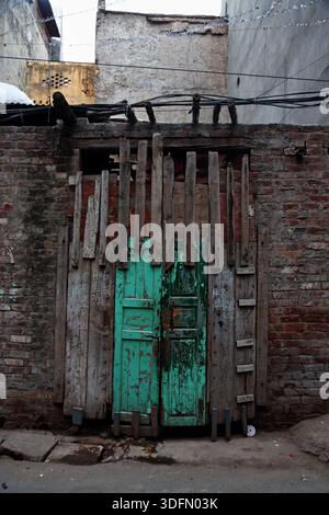 View of an old, weathered green door framed by decaying wooden planks set against a brick wall in a narrow street, Lahore, Punjab, Pakistan. Stock Photo