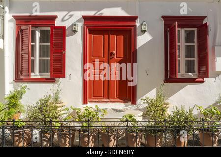 View of the bright red door and window frames contrasting against the stark white walls, framed by potted plants creating a vibrant scene, Mykonos, Gr Stock Photo
