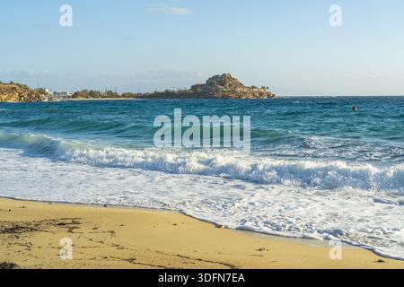 View of turquoise waves crash against a sandy shore, with a rocky outcrop in the distance under a clear blue sky, Mykonos, Mykonos, Greece. Stock Photo
