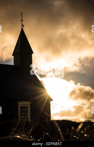 View of the stark silhouette of Budakirkja church piercing the Icelandic skyline as the sun's rays burst through dramatic clouds, Budir, Snaefellsness Stock Photo
