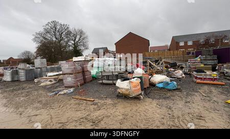 A construction site with stacks of building materials, including bricks and concrete blocks, under a cloudy sky. Muddy ground and trees are visible. Stock Photo