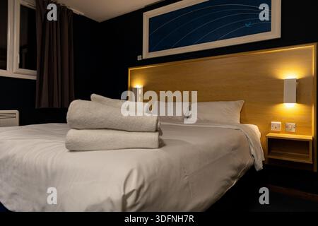 A hotel room features a made bed with white linens and stacked white towels, a wooden headboard with bedside lamps, and artwork on a dark blue wall. Stock Photo