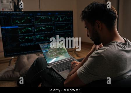 Pensive focused trader male engrossed in financial analysis and day trading, using laptop and large external monitor displaying various stock market Stock Photo