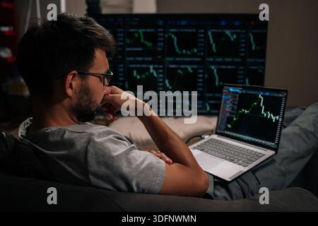 Thoughtful trader male in glasses focused on monitoring complex financial market data and cryptocurrency trends on laptop and large multi-screen setup Stock Photo
