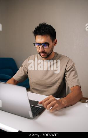 Bearded young businessman wearing glasses concentrating on work on laptop, typing on keyboard in remote home office setting during evening. Stock Photo