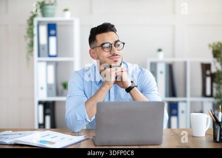 Dreamy Arab guy in glasses sitting at laptop at office, looking aside, taking break from online job Stock Photo