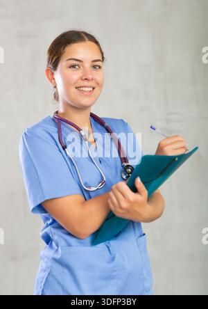 Positive young woman general practitioner in blue uniform filling out clipboard with medical records. Studio photoshoot on gray background Stock Photo