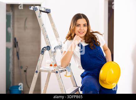 Smiling female builder in blue overalls and yellow hard hat at construction site Stock Photo
