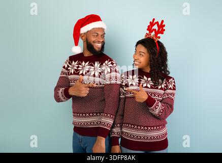 New Year 2022 celebration. Young black couple in Christmas sweaters pointing index fingers at each other Stock Photo