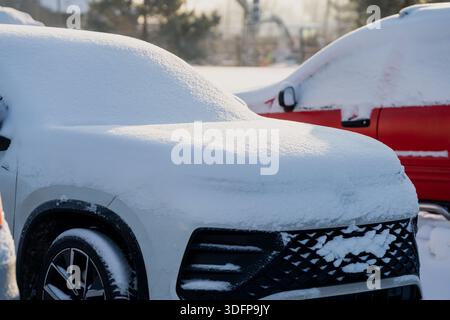Parked SUV completely covered in fresh snow, front grille and wheel partly visible, winter parking lot scene with red snow covered car blurred in back Stock Photo