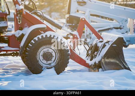 Compact construction loader resting in fresh snow, close view of wheel, hydraulic arms and bucket with golden winter light and machinery yard atmosphe Stock Photo