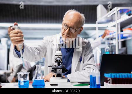 Senior biochemist in lab adding drops of liquid into blue solution to refine new synthetic materials. Elderly man in laboratory mixing chemicals in test tube to watch for reaction Stock Photo