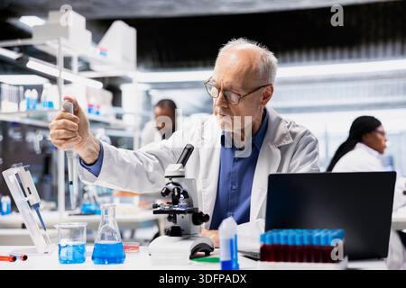 Senior lab manager pipetting agent into blue test tube sample to improve chemical formulation efficiency. Elderly man observing reaction in test tube to verify scientific hypothesis Stock Photo
