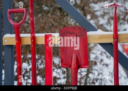 Red metal snow shovels and ice tools hanging on wooden rack outdoors with light snow showing winter maintenance equipment storage and seasonal work to Stock Photo