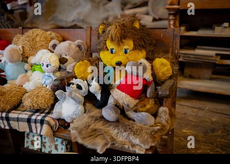 Collection of soft stuffed animals arranged on old wooden seat, teddy bears, lion and small toy figures in warm indoor workshop setting with cluttered Stock Photo
