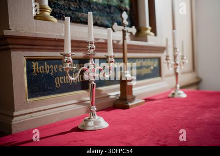 Church altar detail with polished metal candelabras and white candles on red fabric, wooden cross and decorative inscription panel behind in warm inte Stock Photo