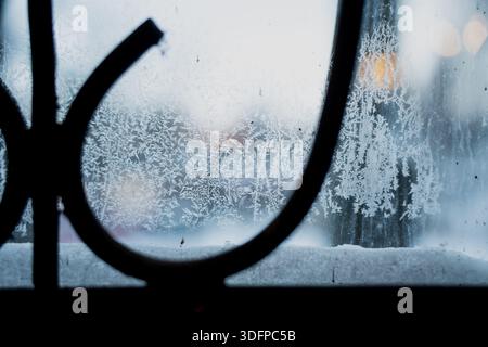 Icy window pane with delicate frost patterns and snow buildup on ledge, shallow depth of field showing cold winter atmosphere with blurred scene outsi Stock Photo