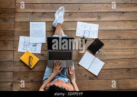 Top view of young lady with laptop, study or work materials and tablet pc sitting on wooden floor indoors, mockup Stock Photo